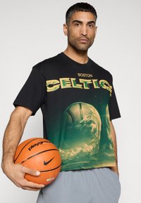 Μαύρο βαμβακερό μπλουζάκι με ζωντανό γραφικό των Boston Celtics, με στρογγυλή λαιμόκοψη και κοντά μανίκια, κρατώντας μια πορτοκαλί μπάλα μπάσκετ.