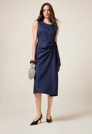 Navy blue, sleeveless dress with a draped design and knot detail at the waist. Paired with a silver beaded clutch and black heeled shoes.
