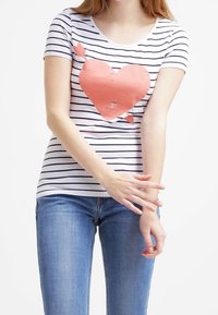 Camiseta blanca de manga corta con rayas horizontales negras y un gráfico de corazón coral desgastado en el centro. Combinada con jeans azules.