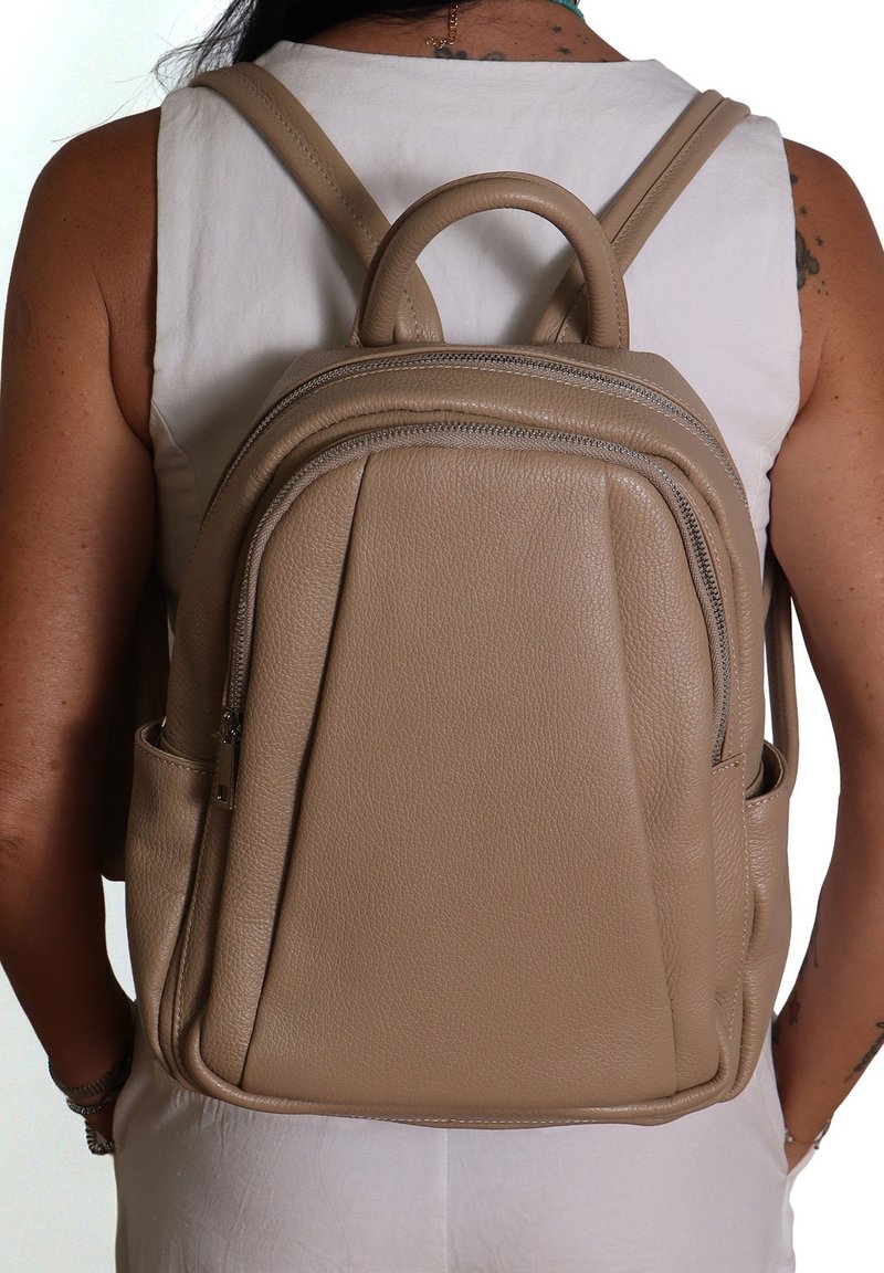 Zaino in pelle beige con doppie tasche con zip, manico superiore arrotondato e spallacci regolabili. Superficie strutturata con accenti minimi.