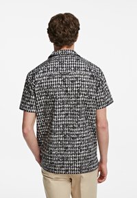 Camicia a maniche corte con un motivo geometrico in bianco e nero. Presenta un colletto classico e un orlo dritto con una vestibilità leggermente sagomata.