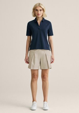 Lyshåret kvinde iført navyblå kortærmet polo, beige plisserede shorts og hvide sneakers, stående mod en ensartet lys baggrund.