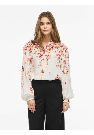 Žena v bielej kvetinovej blúzke s dlhým rukávom s červenými a oranžovými kvetmi, v kombinácii s čiernymi nohavicami s vysokým pásom, stojaca na jednoduchom pozadí.