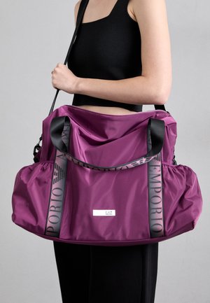 Persona in t-shirt senza maniche nera che tiene una grande borsa a spalla Emporio viola con cinturini neri brandizzati su sfondo grigio.