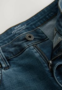 Jean en denim bleu avec une coupe droite, fermeture éclair en métal, boutonnage et étiquette de logo. Décoloration légère et coutures contrastées visibles.