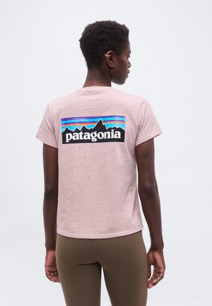 Άτομο με κοντά μαλλιά που φοράει ανοιχτό ροζ μπλούζα Patagonia και καφέ κολάν, εμφανίζεται από πίσω με λευκό φόντο.