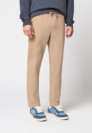 Uomo che indossa pantaloni beige con cordoncino, felpa blu navy e scarpe da ginnastica blu, in piedi contro uno sfondo semplice.