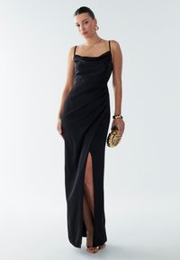 Black evening gown with a cowl neckline, slim fit, side slit, and draped detailing. The model holds a gold-accented clutch and wears statement earrings.
