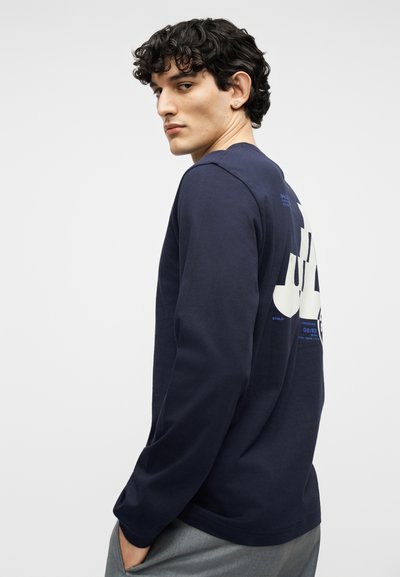 Νεαρός άνδρας με σγουρά μαλλιά, ντυμένος με μπλε σκούρο μακρυμάνικο πουκάμισο που έχει λευκό και μπλε γραφικό κείμενο στο πίσω μέρος, ποζάροντας με τα χέρια στις τσέπες.