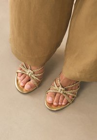 Sandales tressées dorées à semelle plate, avec des brides tissées et des orteils découverts. Les brides forment un motif croisé sur le pied.