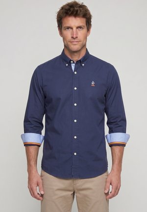 Hombre con camisa azul marino abotonada con puños azul claro remangados y pantalones beige, de pie frente a un fondo gris liso.