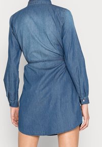 Jeansklänning i medelblått; långa ärmar med knappmanschetter, bältesmidja och rundad nederkant i midjan. Känns slät.