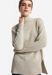 Beige Strickpullover mit hohem Rippkragen und geripptem Saum. Die überschnittenen Schultern und die seitlichen Schlitze sorgen für eine entspannte Passform und Textur.