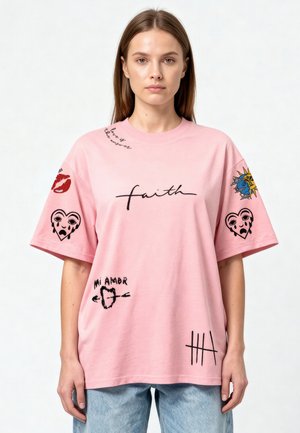 Femme portant un T-shirt rose ample avec des textes noirs et rouges et des motifs graphiques, incluant "faith", des cœurs, le soleil et la lune, et "Mi Amor."