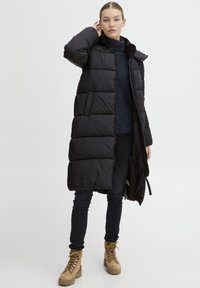 Svart bobledress med foret hette, med kviltet design, sidelommer og åpen front. Modellen har på seg mørke jeans og brune støvler.