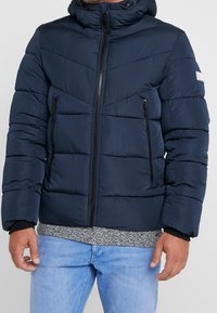 Donkerblauwe donzen jas met capuchon, ritssluitingen en twee zijzakken. Heeft een doorgestikt ontwerp met horizontale en diagonale stiksels.