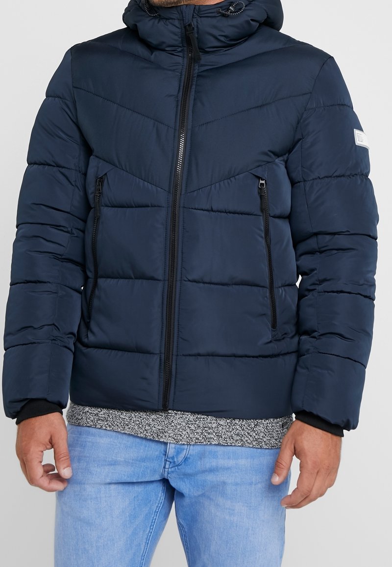 Donkerblauwe donzen jas met capuchon, ritssluitingen en twee zijzakken. Heeft een doorgestikt ontwerp met horizontale en diagonale stiksels.