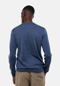 Un homme avec les cheveux courts portant un pull bleu à manches longues et un pantalon beige, vu de dos contre un fond clair.