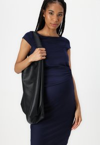 Bolsa tote de couro preto com um acabamento texturizado, design solto e alça larga, segurada por uma pessoa vestindo um vestido azul-marinho.
