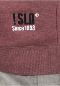 Maroon sweatshirt featuring embroidered text "!SLD © Since 1993" in black and white. Ribbed cuffs and waist add texture.