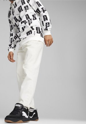 Weißer Hoodie mit großem schwarzem Logodruck, kombiniert mit weißen Hosen und schwarzen Sneakers mit weißen Logoakzenten und Gummisohlen.