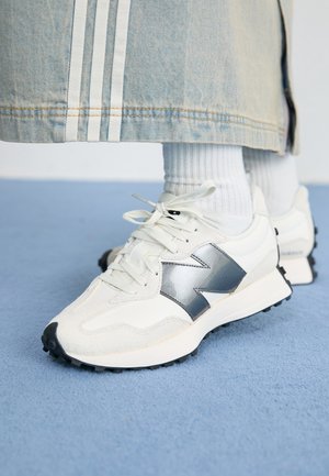 Hvide sneakers med stort sølvlogo brugt med hvide sokker og et lyst denimskørt med hvide striber, stående på en blå gulv.