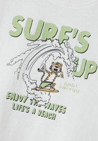Camiseta blanca con un tigre de dibujos animados surfeando una ola, con texto verde: "SURF'S UP", "DISFRUTA LAS OLAS" y "LA VIDA ES UNA PLAYA".