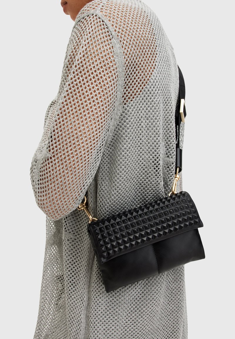 Svart lærveske med teksturert, naglet design, med avtakbar stropp og detaljer i gulltone, båret over en grå mesh-topp.