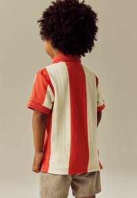 Kortærmet polo skjorte med røde og cremefarvede lodrette striber, tekstureret stof og krave, parret med beige shorts.