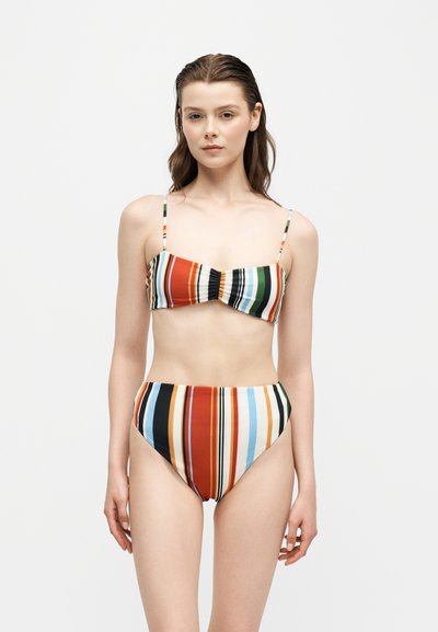Jeune femme debout portant un bikini rayé vertical multicolore avec un haut bandeau et un bas taille haute sur un fond blanc uni.