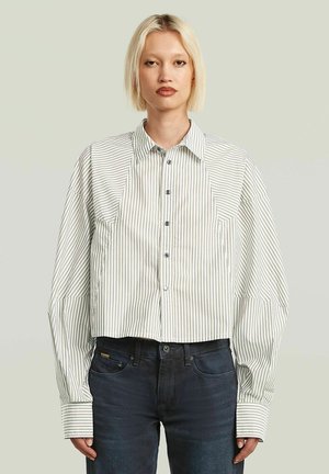 Jeune personne aux cheveux blonds courts porte une chemise blanche ample avec de fines rayures verticales grises et un jean foncé, debout devant un fond uni.