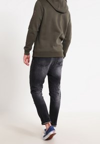 Felpa verde oliva con una patch testurizzato sulla manica sinistra, abbinata a jeans scuri e sneakers blu, vista da dietro.