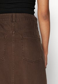 Falda de mezclilla marrón con cintura alta, dos bolsillos traseros y detalles de costura visibles. Textura suave, ligero brillo y diseño ajustado.