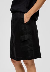 Jupe noire en lin avec une coupe décontractée, poche latérale et ourlet se terminant au-dessus du genou. Le tissu texturé crée un léger éclat.