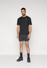 Μαύρο αθλητικό T-shirt με κοντά μανίκια και λογότυπο, σε συνδυασμό με σκούρο σορτς, μαύρες κάλτσες στον αστράγαλο και αθλητικά παπούτσια, σε ουδέτερο φόντο.