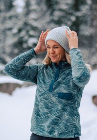 Pull en polaire à motifs bleu clair avec un col boutonné et une poche accentuée, porté avec un bonnet en tricot gris clair devant un paysage enneigé.