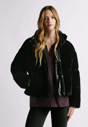 Giubbotto imbottito nero con finitura lucida, texture trapuntata e cappuccio. Presenta chiusure a pressione, tasche e indossato sopra una maglia a righe.