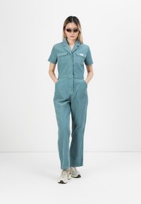 Kvinne iført tealfarget kortermet jumpsuit med frontlommer, beige sneakers og kattøye-solbriller, stående mot en helt hvit bakgrunn.