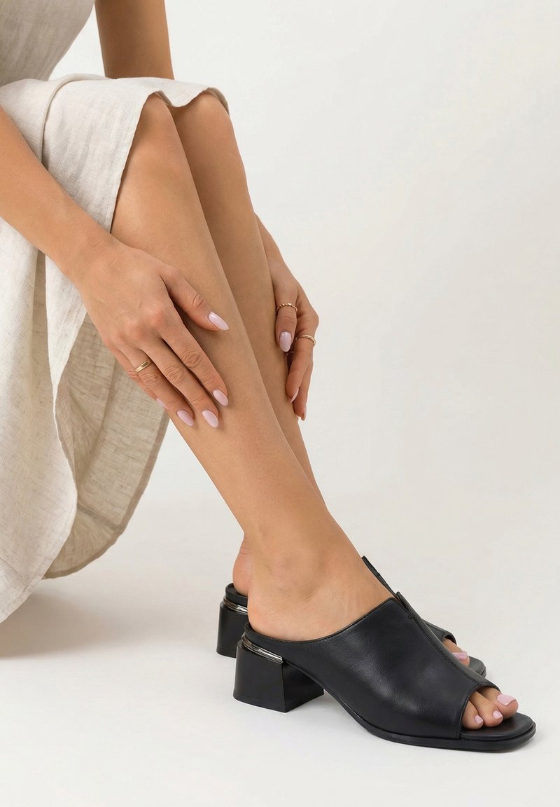 Žena v lehkých lněných šatech, která má na nohou černé otevřené blokové pantofle na podpatku, ruce spočívají na hladkých nohou s bledě růžovým lakem na nehtech.