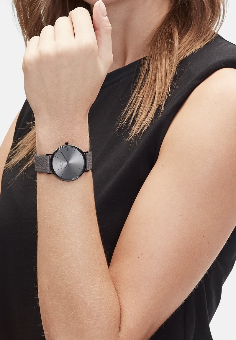Femme portant un haut noir sans manches montre son poignet avec une montre gris foncé au cadran rond minimaliste et au bracelet en cuir.
