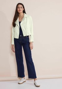 Veste en cuir vert clair avec fermeture éclair et poches latérales, associée à un jean évasé bleu foncé et des baskets blanches sur un fond neutre.