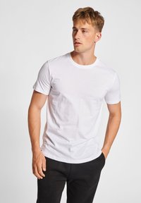 Hvid kortærmet t-shirt i blødt bomuldsstof med rund hals, båret med sorte bukser. Modellen står med hænderne i lommerne.