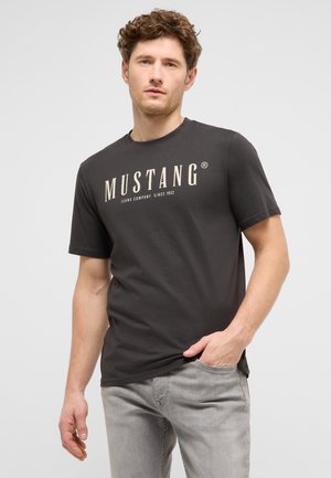 Junger Mann mit lockigem Haar, der ein schwarzes Mustang-T-Shirt und hellgraue Jeans trägt, posiert lässig mit einer Hand in der Tasche.