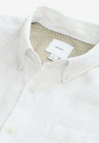 Weißes Leinenhemd mit klassischem Kragen und einer einzelnen Brusttasche. Innen befindet sich ein mit goldfarbenem Muster bedrucktes Etikett mit Größen- und Markeninformationen.
