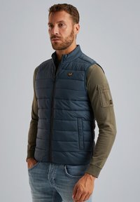 Marinblå pufferväst med horisontell quiltad design, framdragkedja och liten logotyp. Bärs över en långärmad olivgrön skjorta, ihop med ljusa jeans.