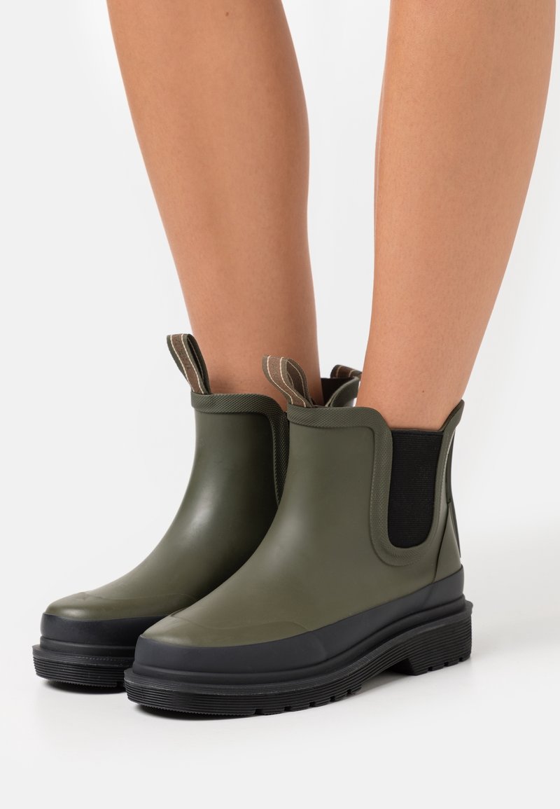 Bottes chevilles en caoutchouc vert olive avec finition mate, panneaux latéraux élastiques et semelles noires texturées. Comprend des languettes de tirage en tissu brun.