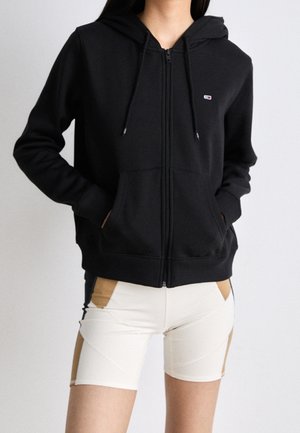 Personne portant un sweat à capuche zippé noir avec les mains dans les poches et un short de vélo en blocs de couleurs beige et marron.