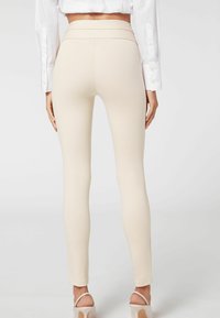 Femme portant des leggings beiges taille haute et une chemise blanche, se tenant avec les bras détendus le long du corps devant un fond uni.
