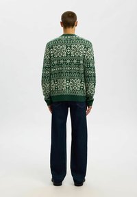 Maglione lavorato a maglia verde con un motivo geometrico bianco, scollatura rotonda e polsini a costine, abbinato a jeans a gamba larga blu scuro.