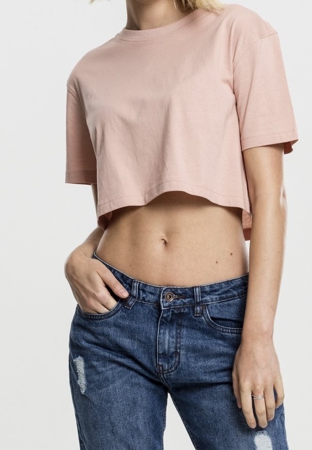 Femme portant un t-shirt court rose clair et un jean bleu déchiré, une main dans la poche et l'autre reposant le long du corps.